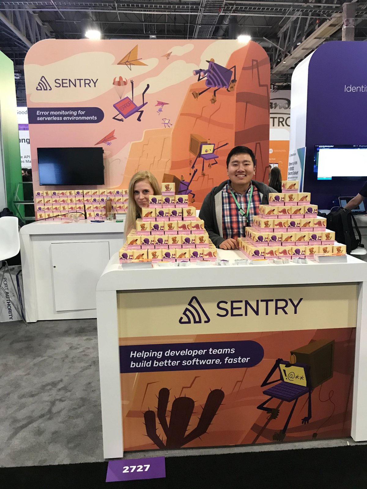Two people behind a Sentry conference booth counter stacked with small boxes, under illustrated panels.