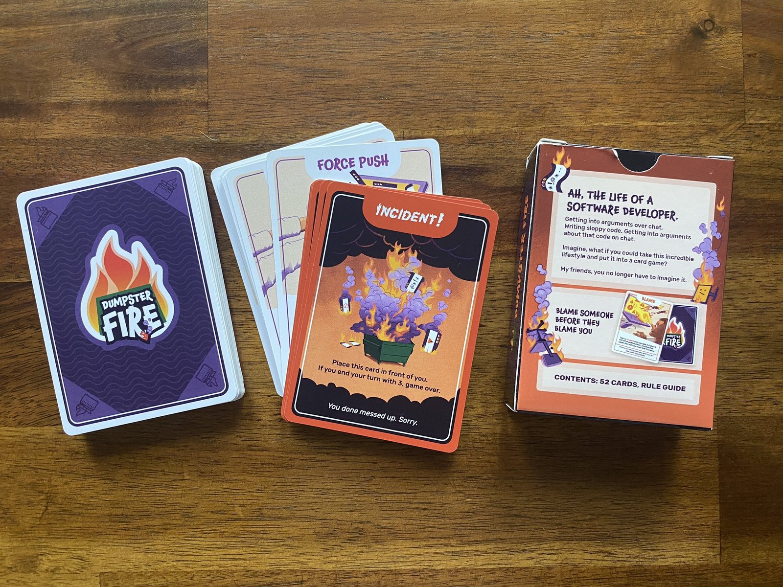 A fan of illustrated Dumpster Fire playing cards beside their box on a wooden table.