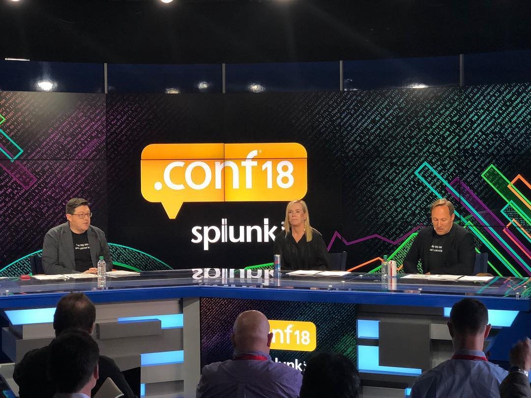 Three people on a broadcast-style set under a .conf18 splunk> sign, in front of colourful angled graphics.